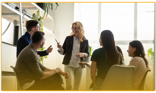 A group of people in an office having a casual meeting with one person standing and gesturing with a pen.