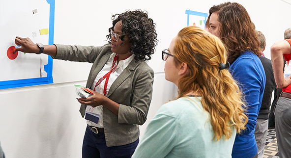 3 women looking at a white board. One is holding a red dot on the  board 